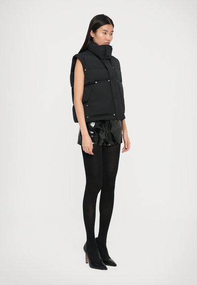 Kvinde iført en sort ærmeløs puffer vest, blanke sorte shorts, sorte strømpebukser og sorte højhælede spidse sko, stående foran en hvid baggrund.
