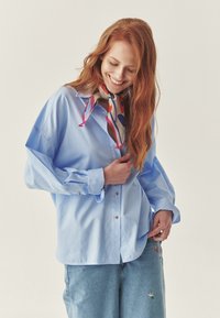Blauw gestreept overhemd met oversized mouwen, gecombineerd met lichte denim en een kleurrijke sjaal met abstracte patronen.