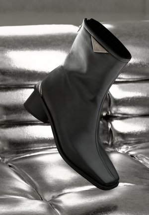 Μαύρο δερμάτινο μποτάκι έως τον αστράγαλο με μυτερή μύτη, μινιμαλιστικό σχέδιο και μικρό τακούνι. Διαθέτει μεταλλικές λεπτομέρειες στο άνω μέρος.