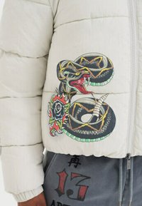 Serpente attorcigliata con la bocca aperta e design di tatuaggio floreale stampato sulla manica di un giubbotto imbottito di colore chiaro.