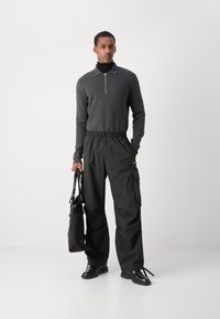 Čierny sveter na zips, voľne strihané čierne cargo nohavice s elastickým pásom a čierne topánky. Model drží čiernu tašku.