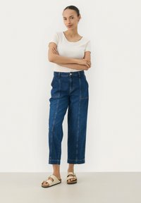 Jeans med hög midja och vida ben, mörkblå, med sömmar framtill och två sidofickor. Matchas med en åtsittande vit topp och beige sandaler.