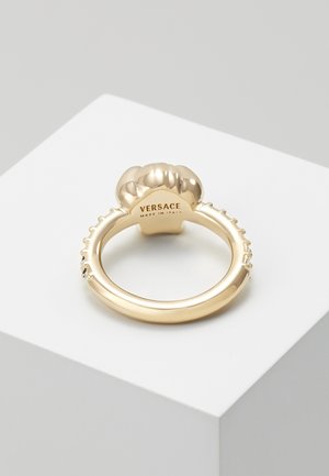 Guld Versace-ring med en rund, skulptureret top og små indlejrede krystaller langs ringen, vist på en hvid pedestal.