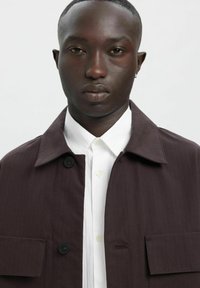 Pruun triibuline jakk nööpidega ja kahe pealise taskuga, kantud valge kraega särgi kohal. Sile tekstuur ja kohandatud disain.