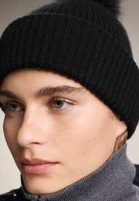 Czarna prążkowana czapka z pomponem na górze, noszona na szarym golfie. Skóra ma gładką teksturę, naturalny odcień i piegi.