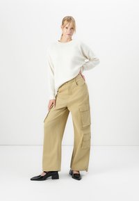 Donna con capelli biondi che indossa un maglione bianco, pantaloni cargo a gamba larga beige e scarpe nere con tacco basso, in piedi su uno sfondo bianco.