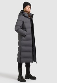 Lange grijze pufferjas met een capuchon, met een gequilte design, zijritsen en gedragen boven een zwarte outfit en zwarte enkellaarsjes.