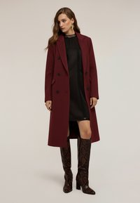 Cappotto borgogna a doppio petto con risvolti, indossato sopra un vestito nero. Presenta stivali al ginocchio con motivo pitone.