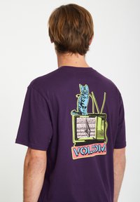 Lila Baumwoll-T-Shirt mit einem grafischen Bild einer Katze auf einem Vintage-Fernseher und stilisiertem Text "VOLCOM" in rosa und blauen Akzenten.