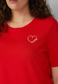 Rotes Baumwoll-T-Shirt mit rundem Halsausschnitt. Mit weißem gesticktem Text und Herzform auf der linken Seite, glatte Textur.