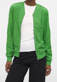 Vihreä pommittaja takki, joka on valmistettu teksturoidusta pitsistä. Takissa on vetoketjusulku, ribbikudoksella varustetut hihansuut ja kaulus, ja se on koristeltu monimutkaisilla kukkakuoseilla.