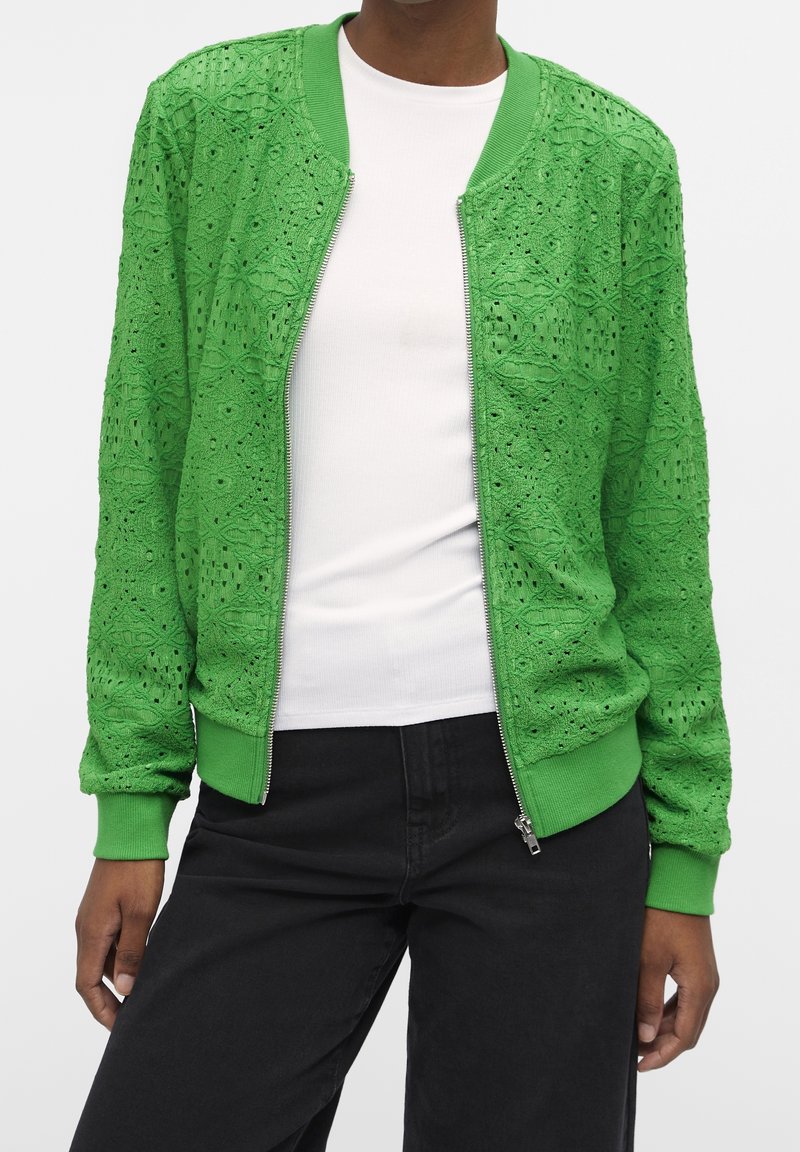 Vihreä pommittaja takki, joka on valmistettu teksturoidusta pitsistä. Takissa on vetoketjusulku, ribbikudoksella varustetut hihansuut ja kaulus, ja se on koristeltu monimutkaisilla kukkakuoseilla.