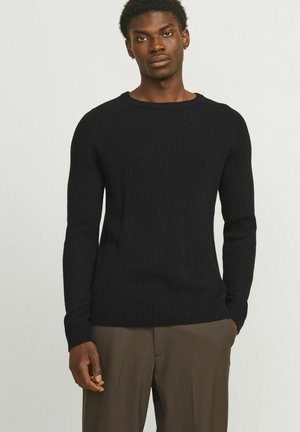 Maglione nero a coste con maniche lunghe, scollatura rotonda e silhouette aderente, abbinato a pantaloni marroni, sfondo in studio.