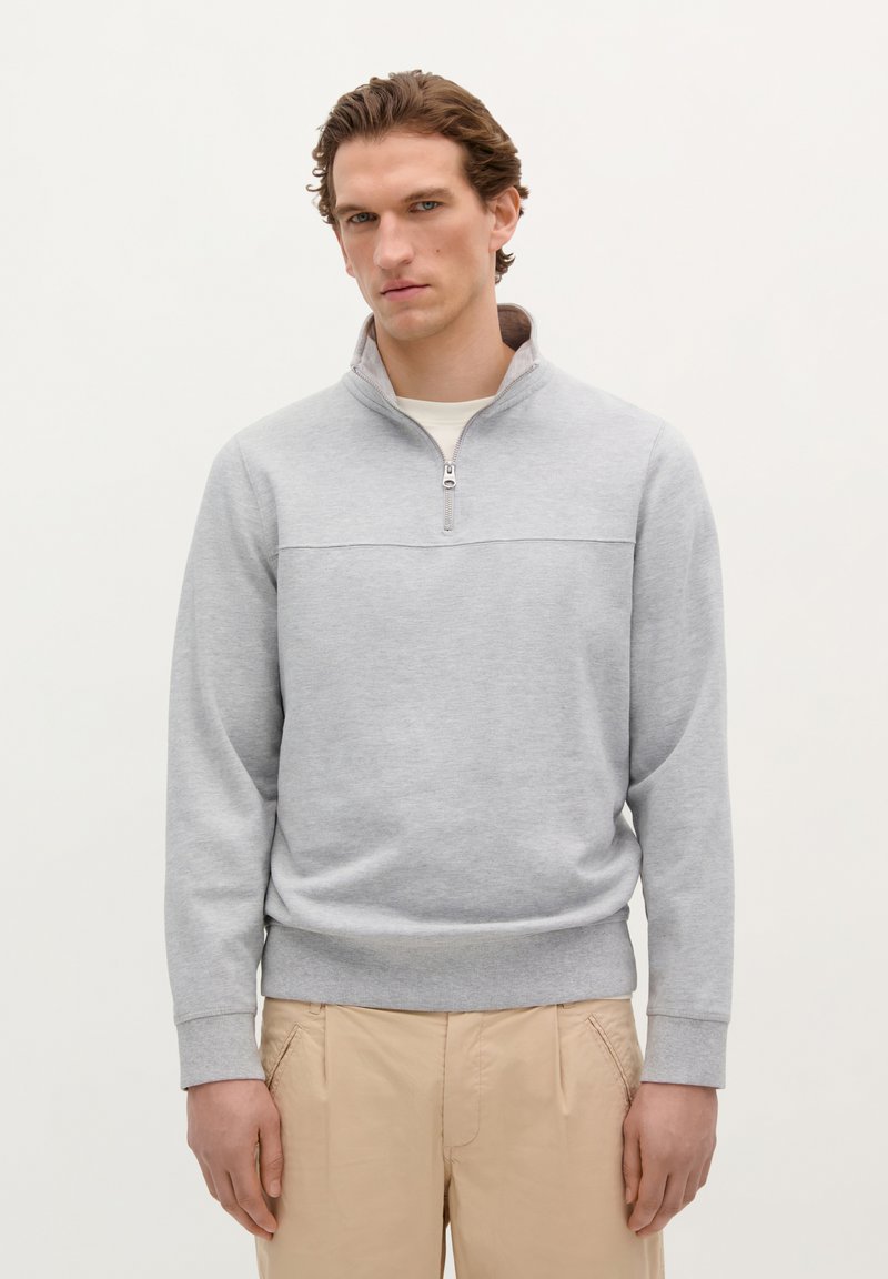 Lichtgrijze ritsvest met een hoge kraag, ribbelmanchetten en onderrand. Draagt over een crèmekleurig shirt, gecombineerd met beige broeken.