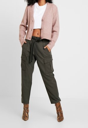 Kvinde iført en kort pink cardigan med knapper, hvid kort tee, olivengrønne cargobukser og brune ankelstøvler i slangeskind, stående med hænderne i lommerne.