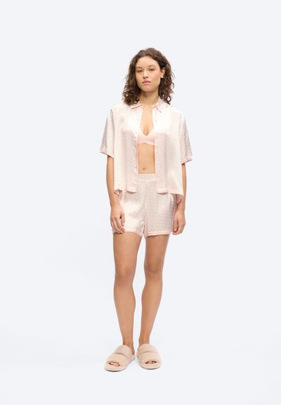 Femme debout portant une chemise en satin rose clair à manches courtes, un short assorti, un soutien-gorge et des chaussons en peluche sur un fond uni.