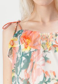 Femme portant une robe à fleurs à épaules dénudées avec des motifs floraux orange, jaune et vert et de fines bretelles nouées.