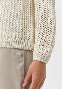 Pull en tricot crème avec un motif en maille ouverte et des poignets côtelés. Présente une forme décontractée et des détails de couture visibles au niveau de l'ourlet.