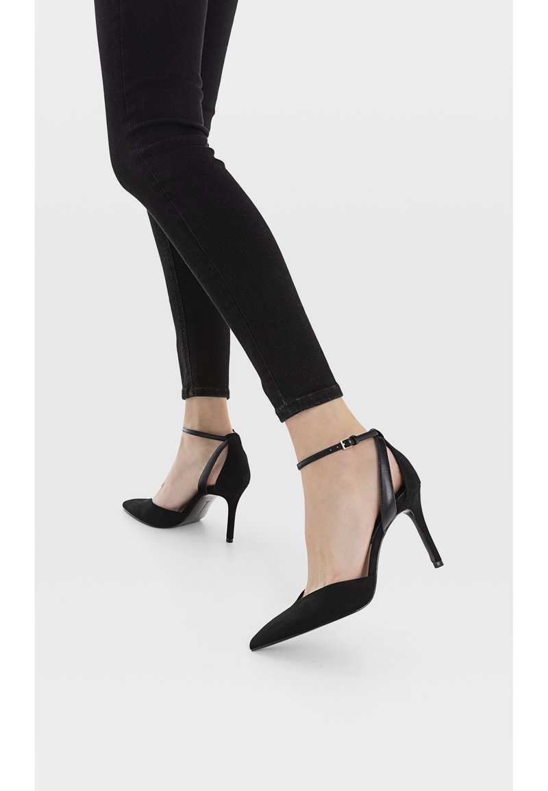 Jambes en jean skinny noir portant des talons hauts noirs à bout pointu avec des brides à la cheville sur fond blanc.