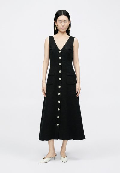 Melna bezroku kleita ar dziļu V-veida izgriezumu, priekšpuses aizdares pogām ar dekoratīviem metāla elementiem un divām kabatām. Gluda tekstūra un A veida siluets.