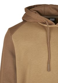 Rudis megztinis su gaubtu, minkštos tekstūros, turintis dviejų tonų dizainą. Gaubtas prigludęs, su raišteliais ir riboto dizaino akcentais ties pečiais.