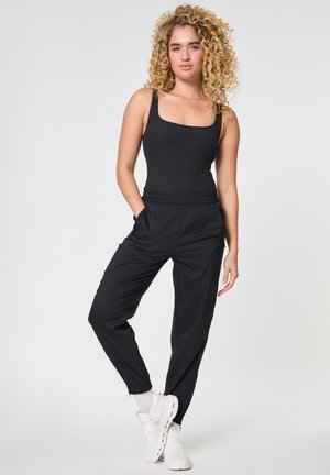 Młoda kobieta z kręconymi blond włosami, ubrana w czarny top, czarne spodnie dresowe, białe skarpetki i białe buty sportowe, stoi z rękami w kieszeniach.