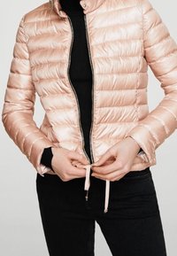 Svetle růžová péřová bunda se zipem vpředu, prošívaným designem, vysokým límcem a elastickými stahovacími šňůrkami na spodním lemu. Není na sobě černý svetr.
