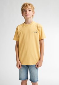 Gul bomulds t-shirt med rund hals, korte ærmer og et lille trykt logo på brystet, parret med lyseblå denim shorts.