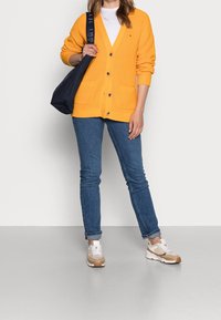 Keltainen ribbineule cardigan, jossa on V-pääntie, puiset napit, kaksi etutaskua, yhdistettynä sinisiin skinny-farkkuihin ja beigeihin lenkkareihin. Musta olkalaukku.