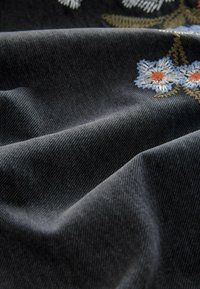 Tissu en denim foncé avec des fleurs brodées bleues et blanches aux centres orange et aux tiges vertes, présenté avec des plis naturels et une texture.