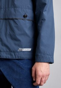 Gros plan d'une personne portant une veste bleue avec un bouton et une étiquette "PS Paul Smith" au-dessus d'un pantalon en velours côtelé bleu.