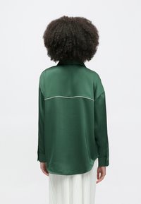 Πράσινο σατινέ πουκάμισο με ελαφρώς άνετη εφαρμογή, με γιακά και αντίθετη λευκή ραφή κατά μήκος της πλάτης. Μακριά μανίκια με μανσέτες.