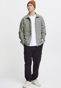 Grijze fleece jas met twee voorzakken, gecombineerd met een wit shirt en zwarte cargobroek. Een bruine gebreide beanie en witte sneakers maken de look compleet.
