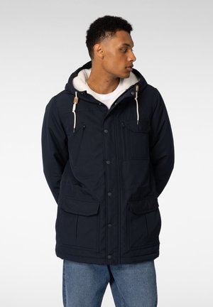 Ung mann vendt sidelengs iført en marineblå parkas med fleeceforet hette, frontlommer og blå jeans mot en ensfarget bakgrunn.