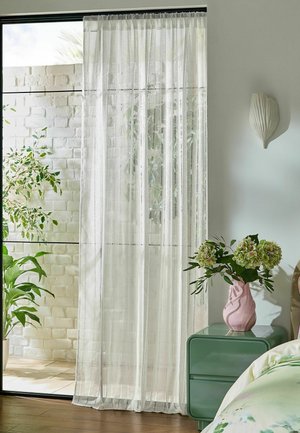 Ein weißer transparenter Vorhang hängt vor einer Glastür mit grünen Pflanzen draußen und einem grünen Nachttisch mit rosa Vase und Blumen drin.