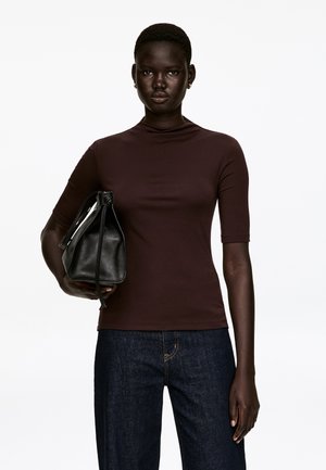 Dark brown, short-sleeve top with a high neckline, paired with dark denim pants and a black leather bag featuring a knotted closure.