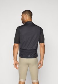 Gilet de cyclisme sans manches noir avec un col haut, un tissu texturé et une poche arrière. Porté sur un short de cyclisme beige.
