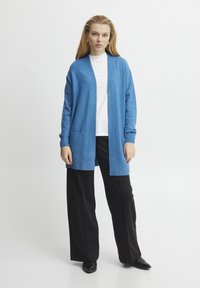 Helleblaue Strickjacke aus weichem Stoff, mit offenem Vorderteil, langen Ärmeln und zwei Fronttaschen, kombiniert mit schwarzen weiten Hosen.