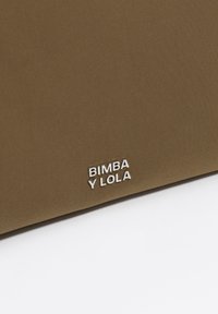 Zbliżenie brązowej tkaniny z metalowymi srebrnymi literami tworzącymi napis "BIMBA Y LOLA" na jasnej krawędzi powierzchni.