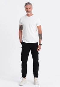 Bílá bavlněná trička v kombinaci s černými cargo kalhotami s bočními kapsami, doplněné bílými sportovními teniskami. Model má krátké šedé vlasy.