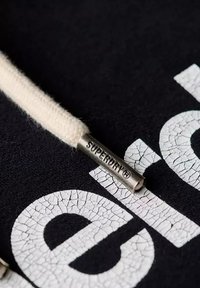 Tela negra con letras blancas agrietadas y un cordón de color crema que presenta un tope de metal plateado grabado con "SUPERDRY."