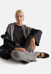 Šedý kardigan přes černý top, šedé texturované kalhoty a černé boty se strapcovým detailem. Černá taška odpočívá vedle. Měkké tkaniny, uvolněný střih.