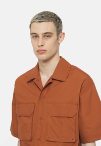 Camisa de manga corta en color óxido, hecha de tejido ligero, presenta dos bolsillos en el pecho, detalles de botones y un cuello puntiagudo.