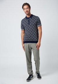 Marinblå polotröja med geometriskt mönster, korta ärmar och ribbade muddar, i kombination med ljusgrå mönstrade byxor och svarta sneakers.