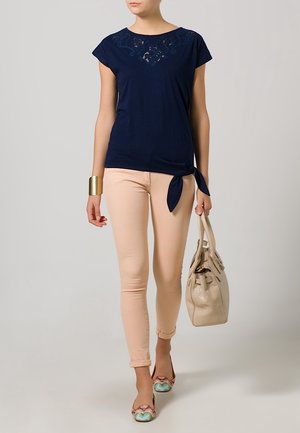 Woman wearing navy lace-accented top with side tie, beige skinny pants, carrying beige handbag, and colorful flat shoes, standing against plain background.