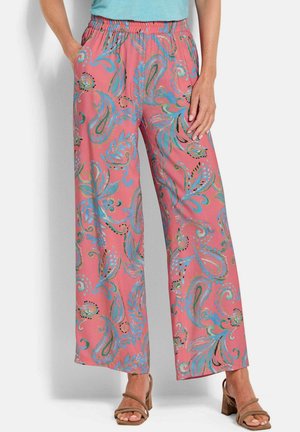 Vrouw die roze wijde broek draagt met blauwgroene paisleypatronen en beige sandalen met hak, handen in de zakken, staand.