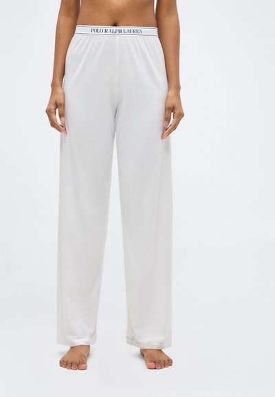 Άτομο που φοράει άνετο άσπρο παντελόνι lounge με ελαστική μέση Polo Ralph Lauren, όρθιο ξυπόλητο μπροστά σε απλό φόντο.