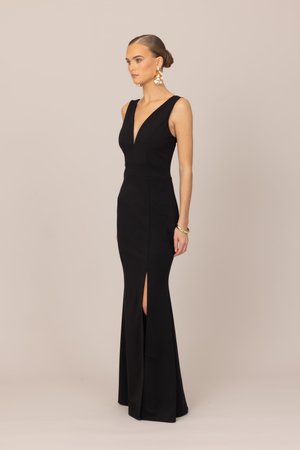 Kobieta stojąca bokiem, ubrana w czarną wieczorową suknię bez rękawów z głębokim dekoltem w serek i rozcięciem z przodu, ozdobiona złotymi kolczykami i bransoletką.