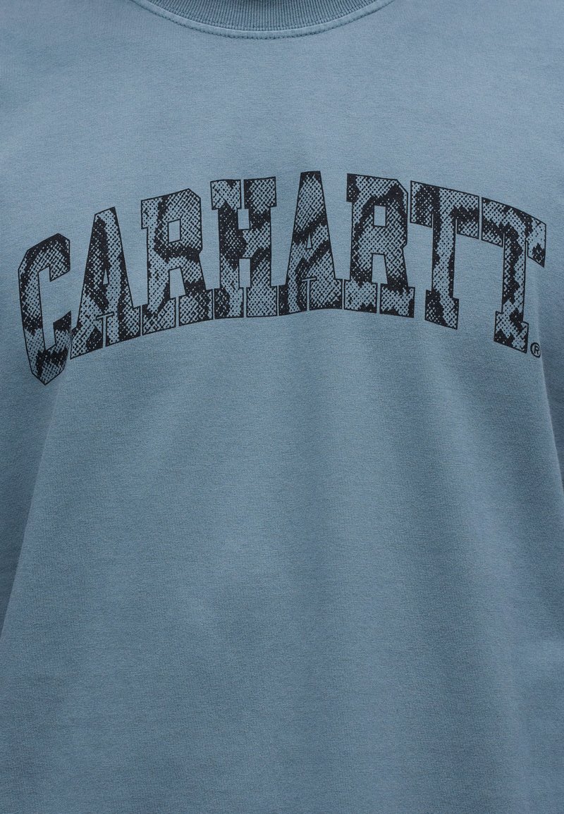 Blauer Stoff mit dem Text „CARHARTT“ in großen, schwarzen, strukturierten Blockbuchstaben, die sich über den Brustbereich biegen.