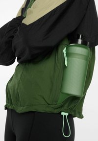 Persoon met een groen en zwart jack en een groene waterfles in een zijzak, tegen een witte achtergrond.